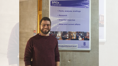 A photo of Dan in front of the Scottish Parliament Information Centre (SPICe) information board