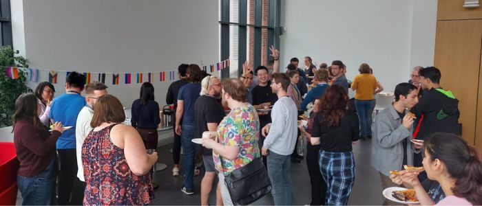 People gathered indoors for a social event, standing with plates of food near tables, under a string of international flags, engaging in conversation and serving themselves.