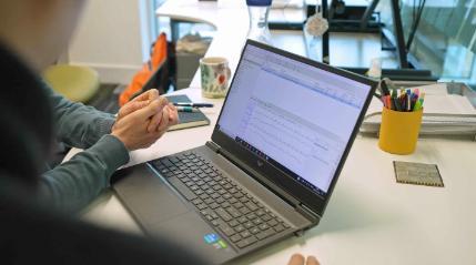 A person sits at a desk with a laptop open to a data analysis program showing several line graphs. Their hands are clasped in front of the keyboard.