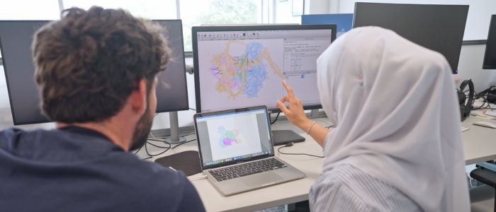 Two people sit at a desk looking at molecular or structural graphics on a laptop and a large monitor, discussing the visual data.
